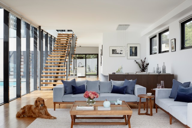 A bright living room with a wooden staircase, modern furniture, and a golden retriever lying on the floor, featuring large windows and a pool view.